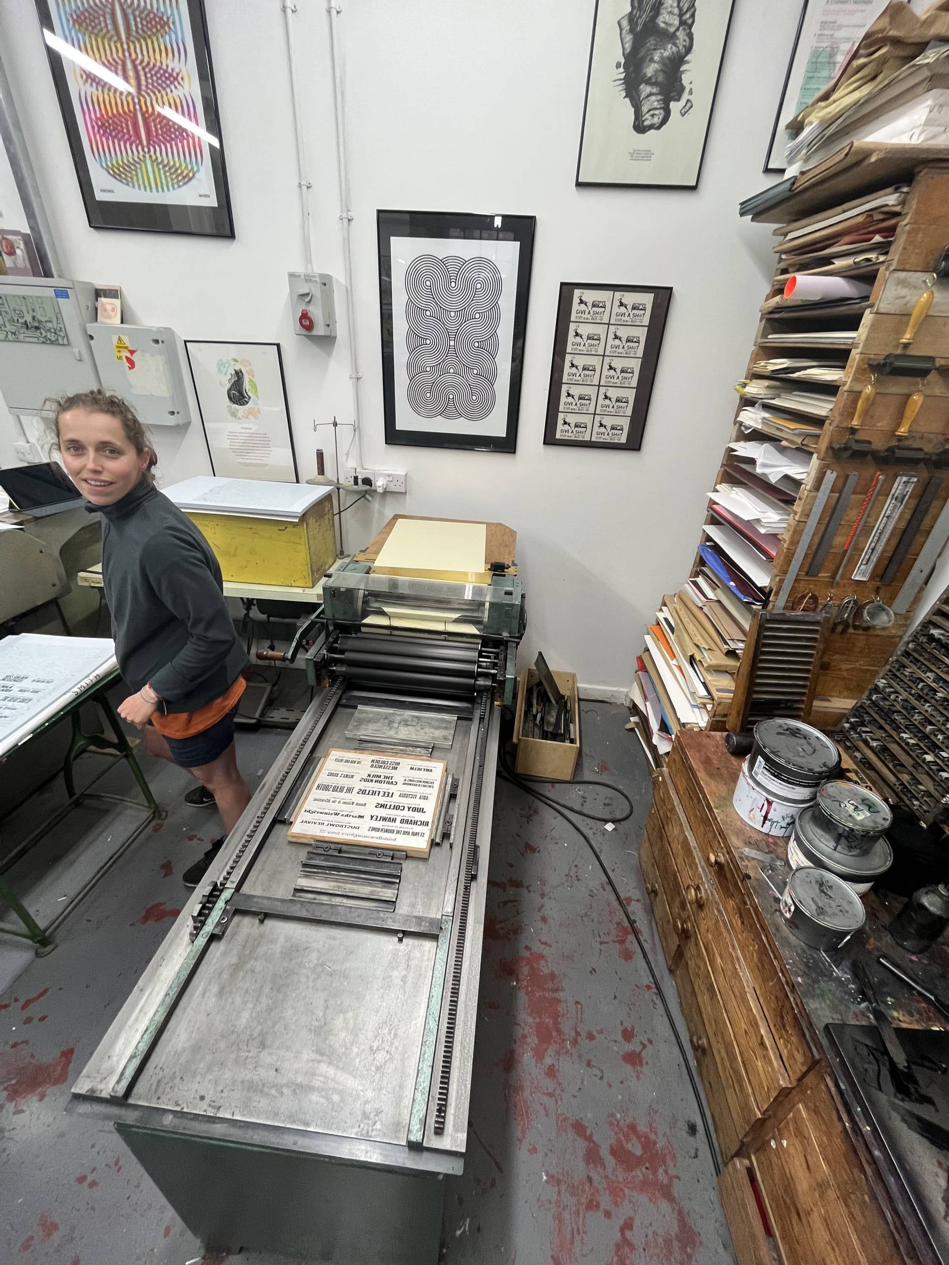 A woman stands next to a large metal printing press surrounded by shelves of inks and letter stamps.